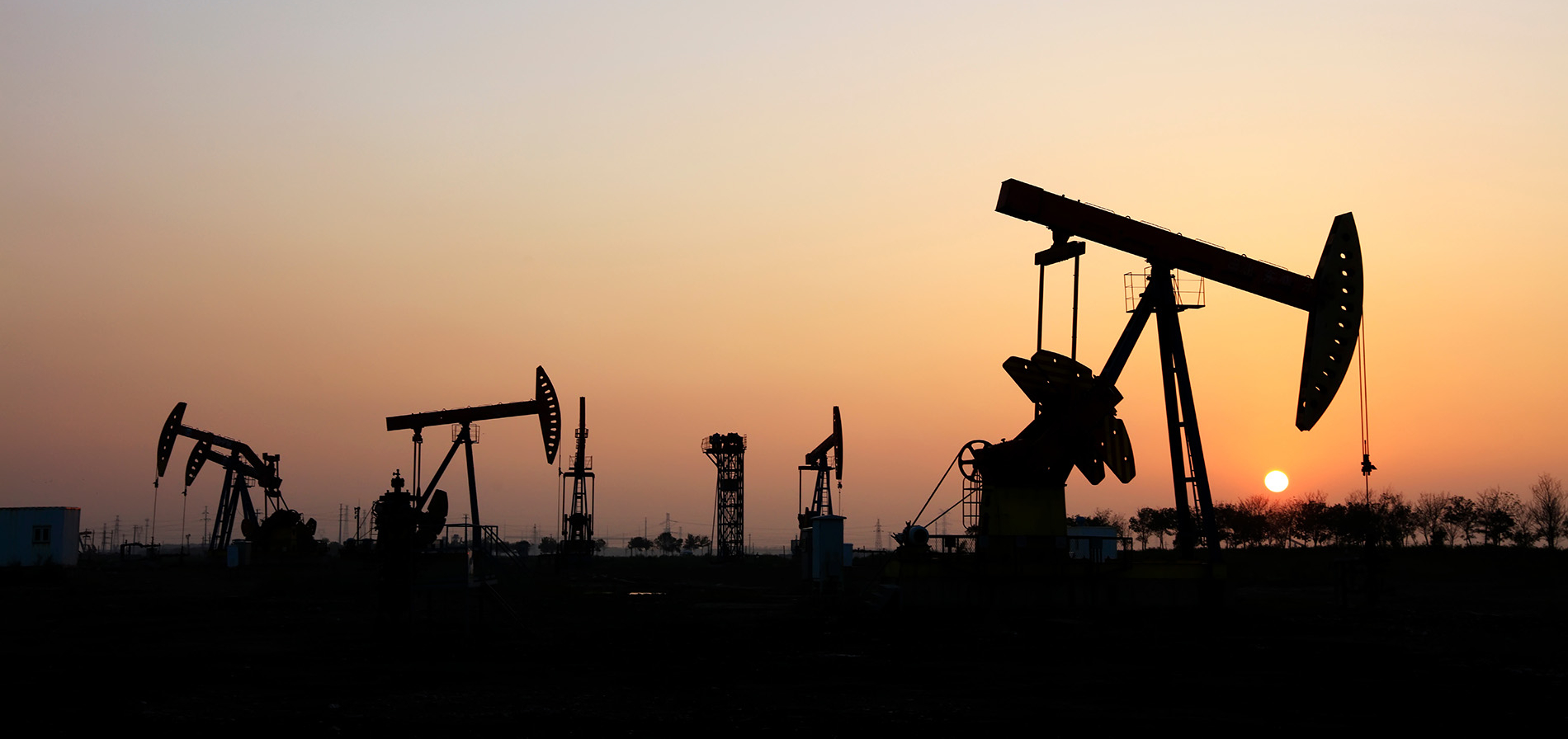 Silhouettes of Oil & Gas pumpjacks and industrial structures stand against an orange sunset sky, with the sun low on the horizon and sparse vegetation in the foreground.
